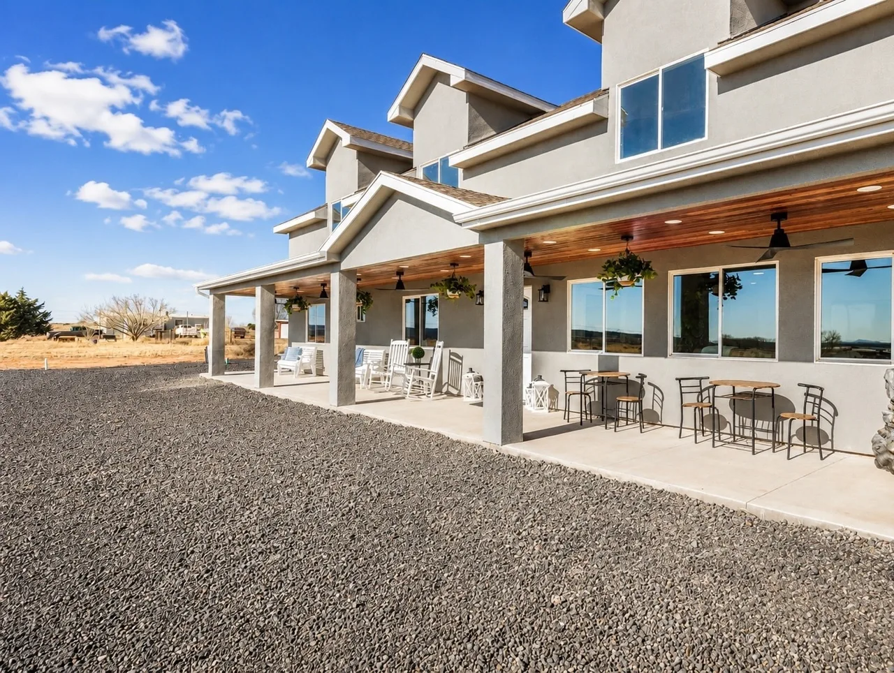 Covered front patio at Zion Luxury Retreat with white rocking chairs, iron pub tables, wood-plank ceiling fans, and stucco columns above a gravel yard.