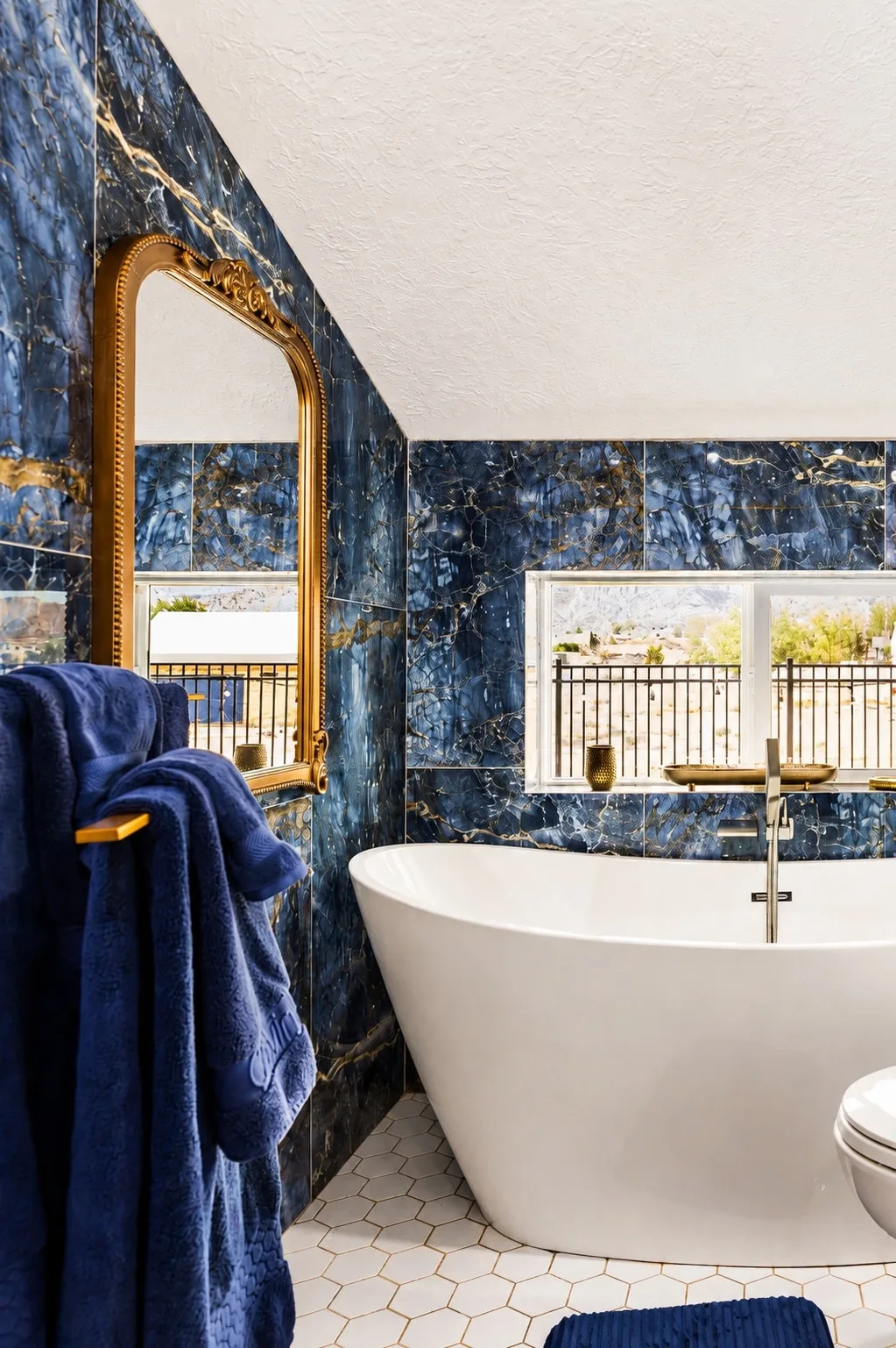 Sky Suite's vaulted primary bathroom clad in dramatic blue-and-gold marble with a white freestanding soaking tub and a small balcony-rail window.